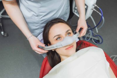 A woman in a dental chair is being prepared for a procedure with a sedation mask placed over her nose.