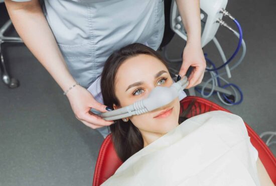 A woman in a dental chair is being prepared for a procedure with a sedation mask placed over her nose.