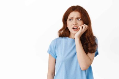 A woman with long red hair in a blue shirt looks anxious, biting her nails and gazing upward against a plain white background.